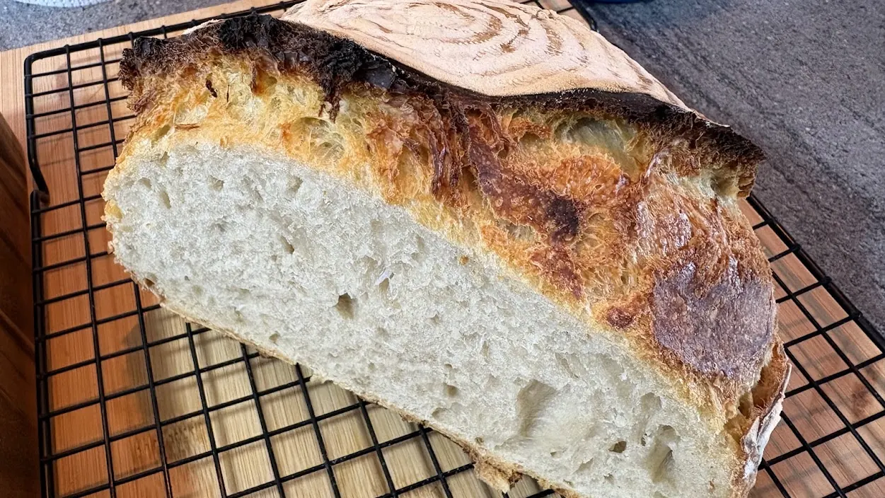 A sourdough loaf on a wire cooling rack, cut open to show an open, airy crumb inside a dark, blistered crust.